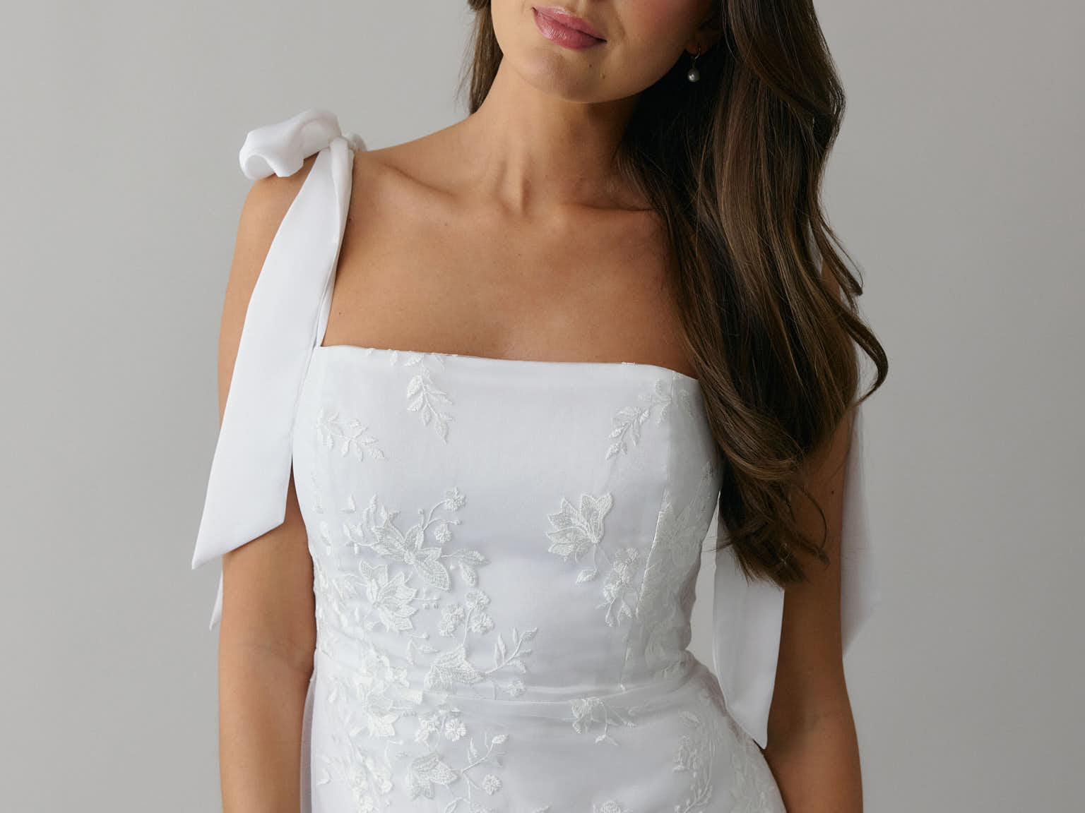 A woman stands against a light grey backdrop. She's wearing a white mini dress with white floral embroidery and satin tie straps. This bridal event dress is called the Tanner Mini.