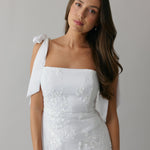 A woman stands against a light grey backdrop. She's wearing a white mini dress with white floral embroidery and satin tie straps. This bridal event dress is called the Tanner Mini.