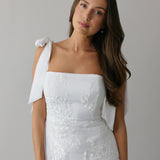 A woman stands against a light grey backdrop. She's wearing a white mini dress with white floral embroidery and satin tie straps. This bridal event dress is called the Tanner Mini.