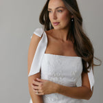 A woman stands against a light grey backdrop. She's wearing a white mini dress with white floral embroidery and satin tie straps. This bridal event dress is called the Tanner Mini.