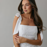 A woman stands against a light grey backdrop. She's wearing a white mini dress with white floral embroidery and satin tie straps. This bridal event dress is called the Tanner Mini.