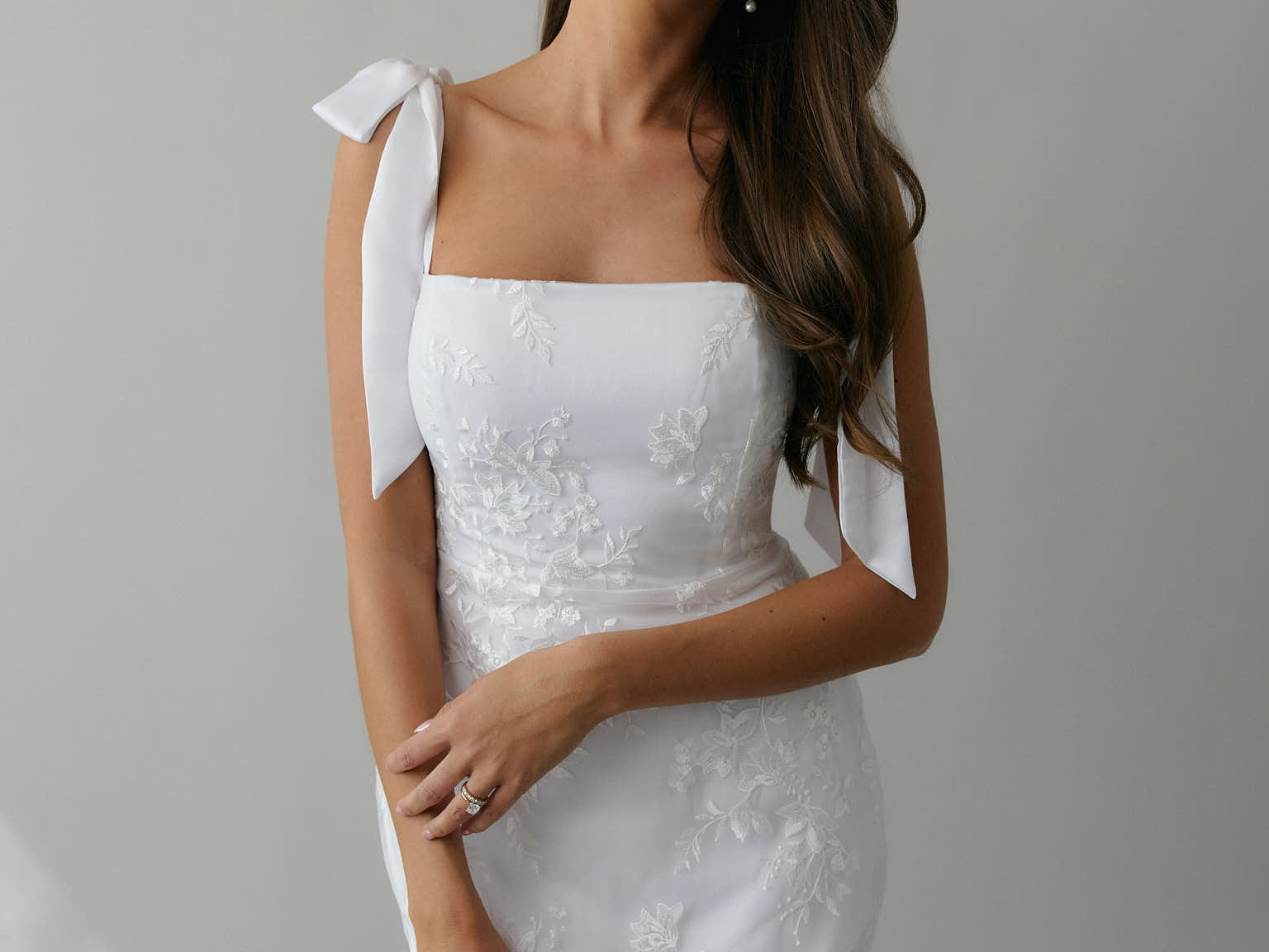 A woman stands against a light grey backdrop. She's wearing a white mini dress with white floral embroidery and satin tie straps. This bridal event dress is called the Tanner Mini.