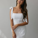 A woman stands against a light grey backdrop. She's wearing a white mini dress with white floral embroidery and satin tie straps. This bridal event dress is called the Tanner Mini.