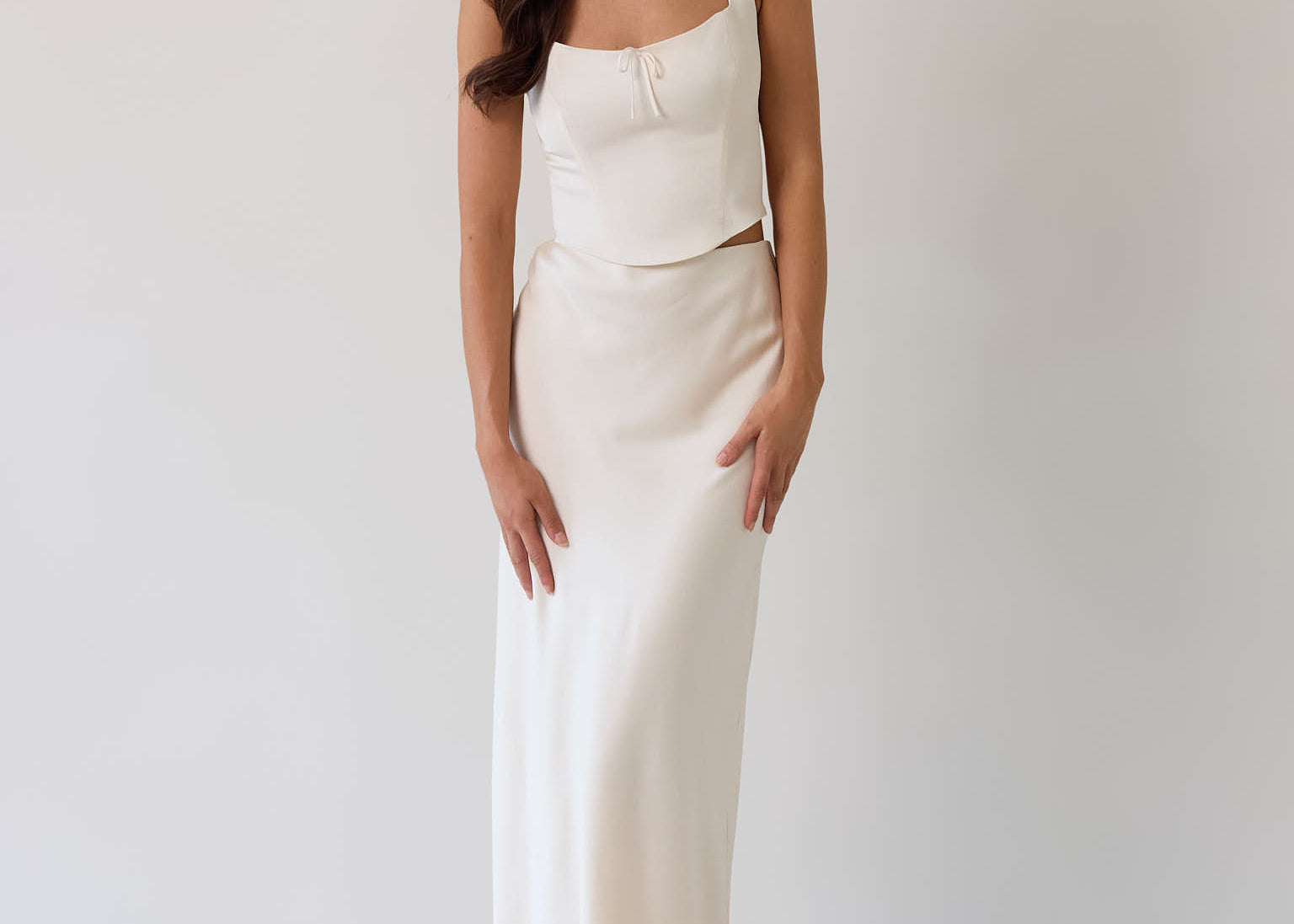 This is the Conrad Set in Ivory. This is a two piece satin set with a fitted, corseted top with thick straps and a delicate tie at the bust and a slight basque waist. It comes with a floor length satin column skirt. 