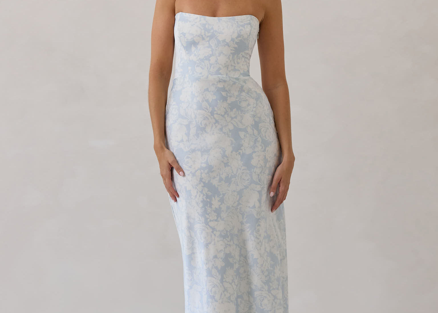 A woman is standing infront of a white backdrop. She's wearing a light blue and white floral print strapless satin dress. The dress is fitted through the bodice and falls nicely over the hips and is tea-length. This dress can be worn as a bridesmaid dress or for any special event. This is the Brodie Dress in Serene. 