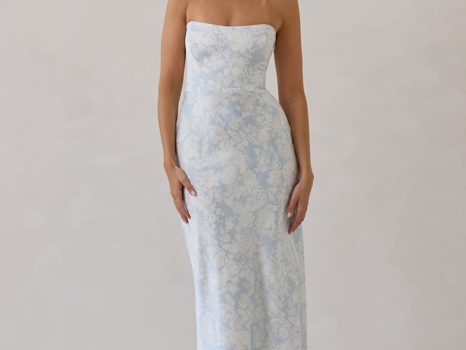 A woman is standing infront of a white backdrop. She's wearing a light blue and white floral print strapless satin dress. The dress is fitted through the bodice and falls nicely over the hips and is tea-length. This dress can be worn as a bridesmaid dress or for any special event. This is the Brodie Dress in Serene. 