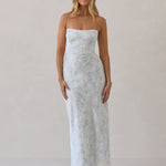 A woman is standing infront of a white backdrop. She's wearing a light blue and white floral print strapless satin dress. The dress is fitted through the bodice and falls nicely over the hips and is tea-length. This dress can be worn as a bridesmaid dress or for any special event. This is the Brodie Dress in Serene. 