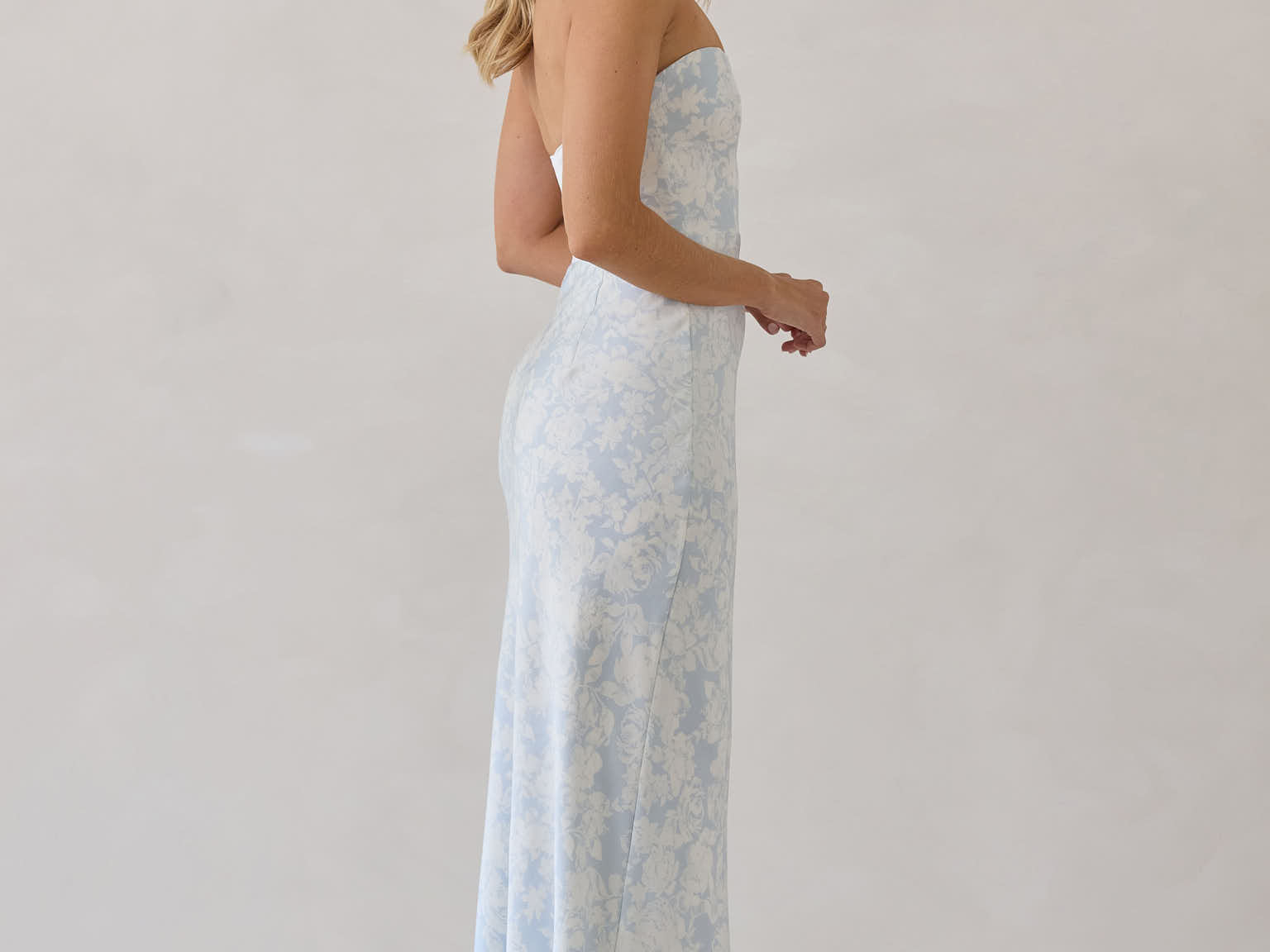 A woman is standing infront of a white backdrop facing the right. She's wearing a light blue and white floral print strapless satin dress. The dress is fitted through the bodice and falls nicely over the hips and is tea-length. This dress can be worn as a bridesmaid dress or for any special event. This is the Brodie Dress in Serene. 