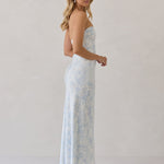 A woman is standing infront of a white backdrop facing the right. She's wearing a light blue and white floral print strapless satin dress. The dress is fitted through the bodice and falls nicely over the hips and is tea-length. This dress can be worn as a bridesmaid dress or for any special event. This is the Brodie Dress in Serene. 