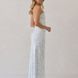 A woman is standing infront of a white backdrop facing the right. She's wearing a light blue and white floral print strapless satin dress. The dress is fitted through the bodice and falls nicely over the hips and is tea-length. This dress can be worn as a bridesmaid dress or for any special event. This is the Brodie Dress in Serene. 