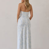 A woman is standing infront of a white backdrop facing the back. She's wearing a light blue and white floral print strapless satin dress. The dress is fitted through the bodice and falls nicely over the hips and is tea-length. This dress can be worn as a bridesmaid dress or for any special event. This is the Brodie Dress in Serene. 
