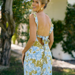 A woman is standing outside wearing a blue and green floral print bridesmaid dress with an open back. The dress features a slight ruffle detail along the strap and slit of the skirt. This is the Avondale Dress in Frankie.