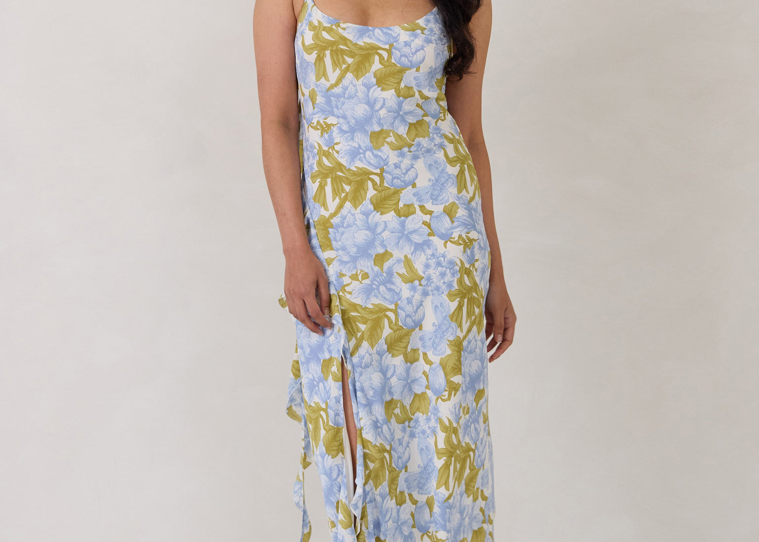 A woman is standing against a white backdrop  wearing a blue and green floral print bridesmaid dress with an open back. The dress features a slight ruffle detail along the strap and slit of the skirt. This is the Avondale Dress in Frankie.