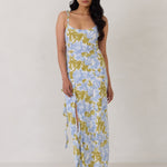 A woman is standing against a white backdrop  wearing a blue and green floral print bridesmaid dress with an open back. The dress features a slight ruffle detail along the strap and slit of the skirt. This is the Avondale Dress in Frankie.