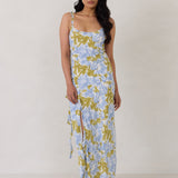 A woman is standing against a white backdrop  wearing a blue and green floral print bridesmaid dress with an open back. The dress features a slight ruffle detail along the strap and slit of the skirt. This is the Avondale Dress in Frankie.