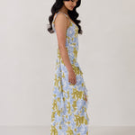 A woman is standing against a white backdrop  wearing a blue and green floral print bridesmaid dress with an open back. The dress features a slight ruffle detail along the strap and slit of the skirt. This is the Avondale Dress in Frankie.
