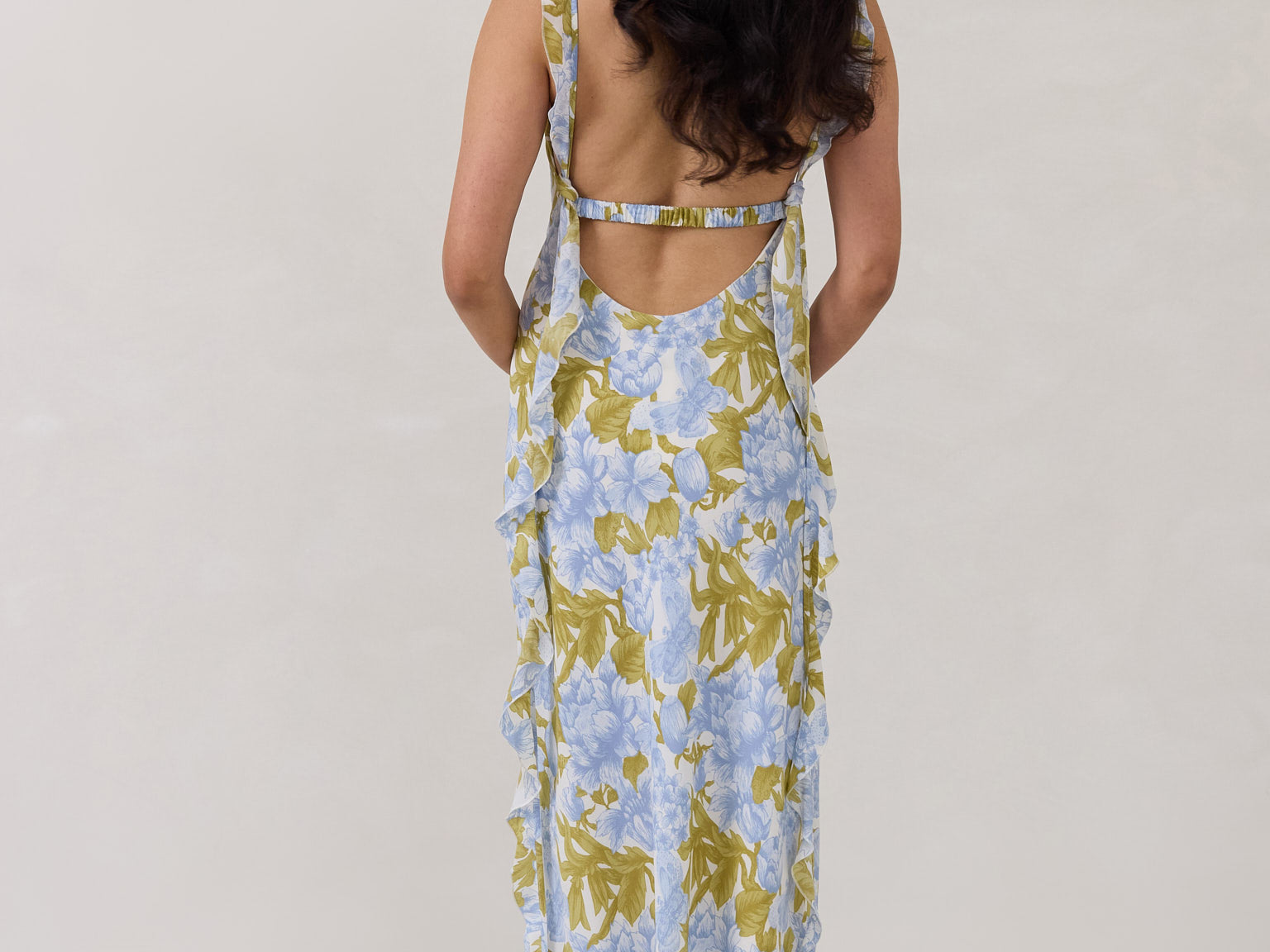 A woman is standing against a white backdrop  wearing a blue and green floral print bridesmaid dress with an open back. The dress features a slight ruffle detail along the strap and slit of the skirt. This is the Avondale Dress in Frankie.