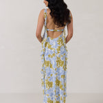 A woman is standing against a white backdrop  wearing a blue and green floral print bridesmaid dress with an open back. The dress features a slight ruffle detail along the strap and slit of the skirt. This is the Avondale Dress in Frankie.