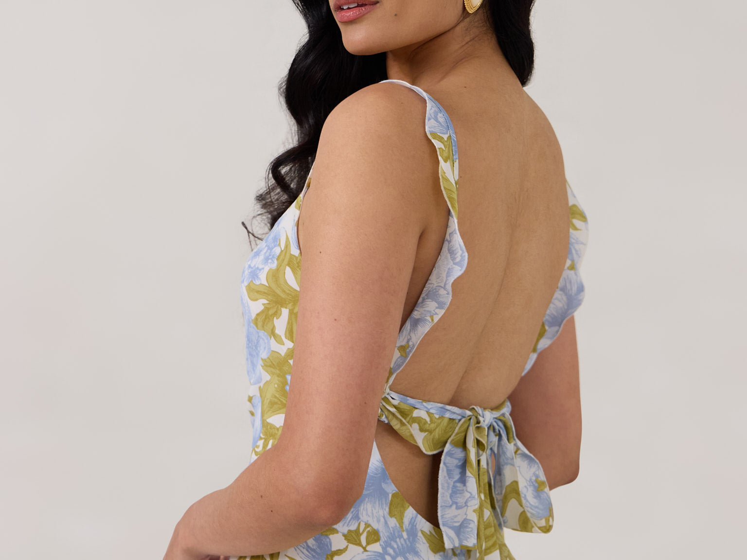 A woman is standing against a white backdrop  wearing a blue and green floral print bridesmaid dress with an open back. The dress features a slight ruffle detail along the strap and slit of the skirt. This is the Avondale Dress in Frankie.