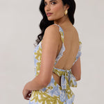 A woman is standing against a white backdrop  wearing a blue and green floral print bridesmaid dress with an open back. The dress features a slight ruffle detail along the strap and slit of the skirt. This is the Avondale Dress in Frankie.