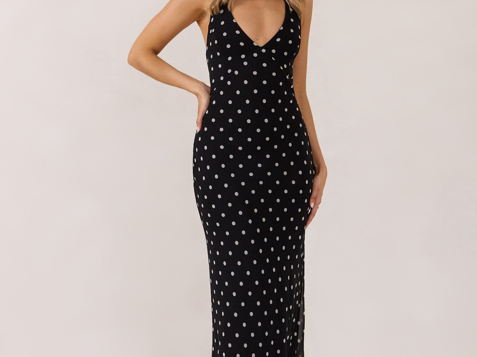 A woman stands against a white backdrop wearing a black and white polka dot dress. The dress has a halter neckline with an optional scarf detail. This is the Archer Dress in Midnight Dot.