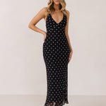 A woman stands against a white backdrop wearing a black and white polka dot dress. The dress has a halter neckline with an optional scarf detail. This is the Archer Dress in Midnight Dot.