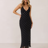 A woman stands against a white backdrop wearing a black and white polka dot dress. The dress has a halter neckline with an optional scarf detail. This is the Archer Dress in Midnight Dot.