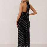 A woman stands against a white backdrop wearing a black and white polka dot dress. The dress has a halter neckline with an optional scarf detail. This is the Archer Dress in Midnight Dot.