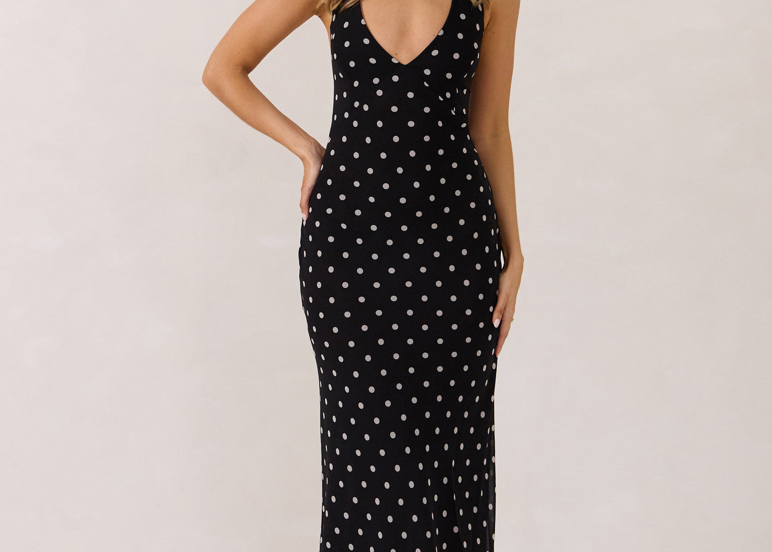 A woman stands against a white backdrop wearing a black and white polka dot dress. The dress has a halter neckline with an optional scarf detail. This is the Archer Dress in Midnight Dot.