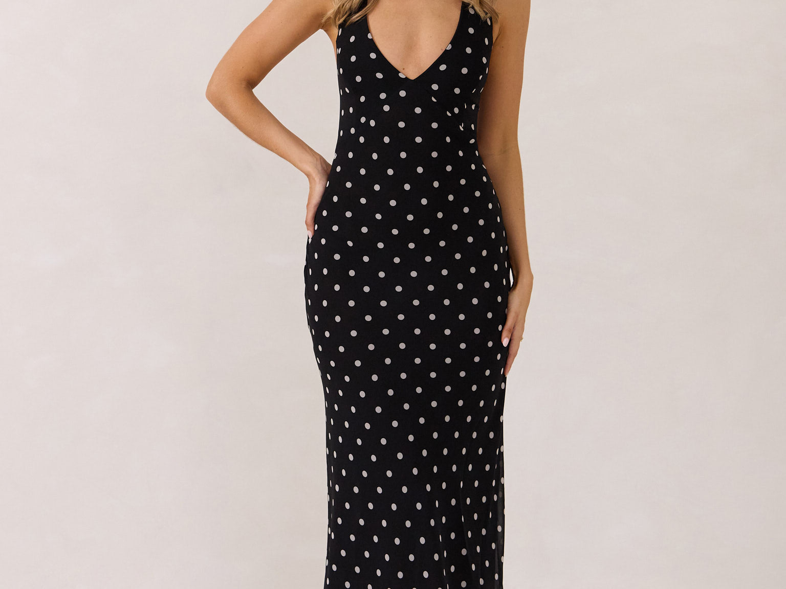 A woman stands against a white backdrop wearing a black and white polka dot dress. The dress has a halter neckline with an optional scarf detail. This is the Archer Dress in Midnight Dot.
