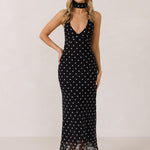 A woman stands against a white backdrop wearing a black and white polka dot dress. The dress has a halter neckline with an optional scarf detail. This is the Archer Dress in Midnight Dot.