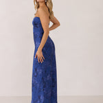 A woman stands against a white backdrop facing the left. She's wearing an indigo blue strapless dress with a blue floral print. The dress is fitted through the bodice, and falls nicely over the hips and is tea-length. This is the Brodie Dress in Azure. 