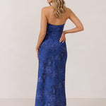 A woman stands against a white backdrop facing the back. She's wearing an indigo blue strapless dress with a blue floral print. The dress is fitted through the bodice, and falls nicely over the hips and is tea-length. This is the Brodie Dress in Azure. 