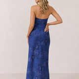 A woman stands against a white backdrop facing the back. She's wearing an indigo blue strapless dress with a blue floral print. The dress is fitted through the bodice, and falls nicely over the hips and is tea-length. This is the Brodie Dress in Azure. 