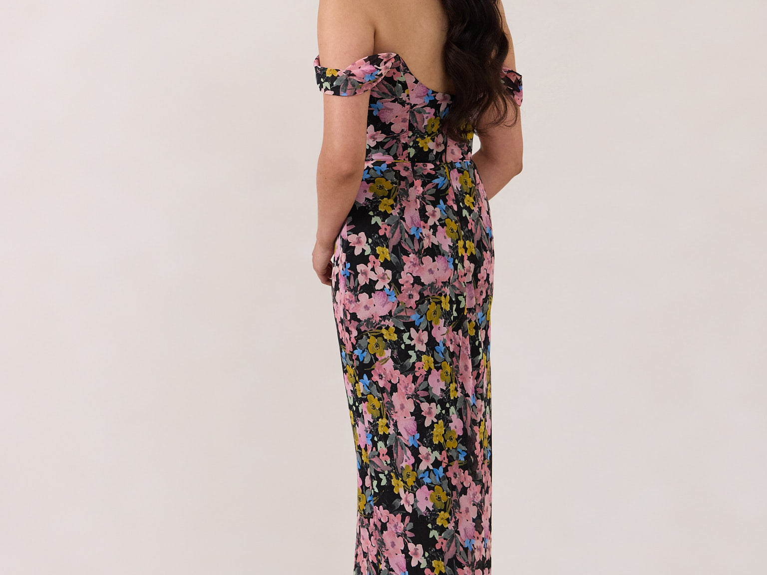 A woman is standing in front of a white background facing the back. She's wearing a bold pink and blue floral print strapless dress with an off-the-shoulder strap. This dress is Sutton in Liora and is perfect as a wedding guest dress, or for any special event. 