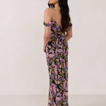 A woman is standing in front of a white background facing the back. She's wearing a bold pink and blue floral print strapless dress with an off-the-shoulder strap. This dress is Sutton in Liora and is perfect as a wedding guest dress, or for any special event. 