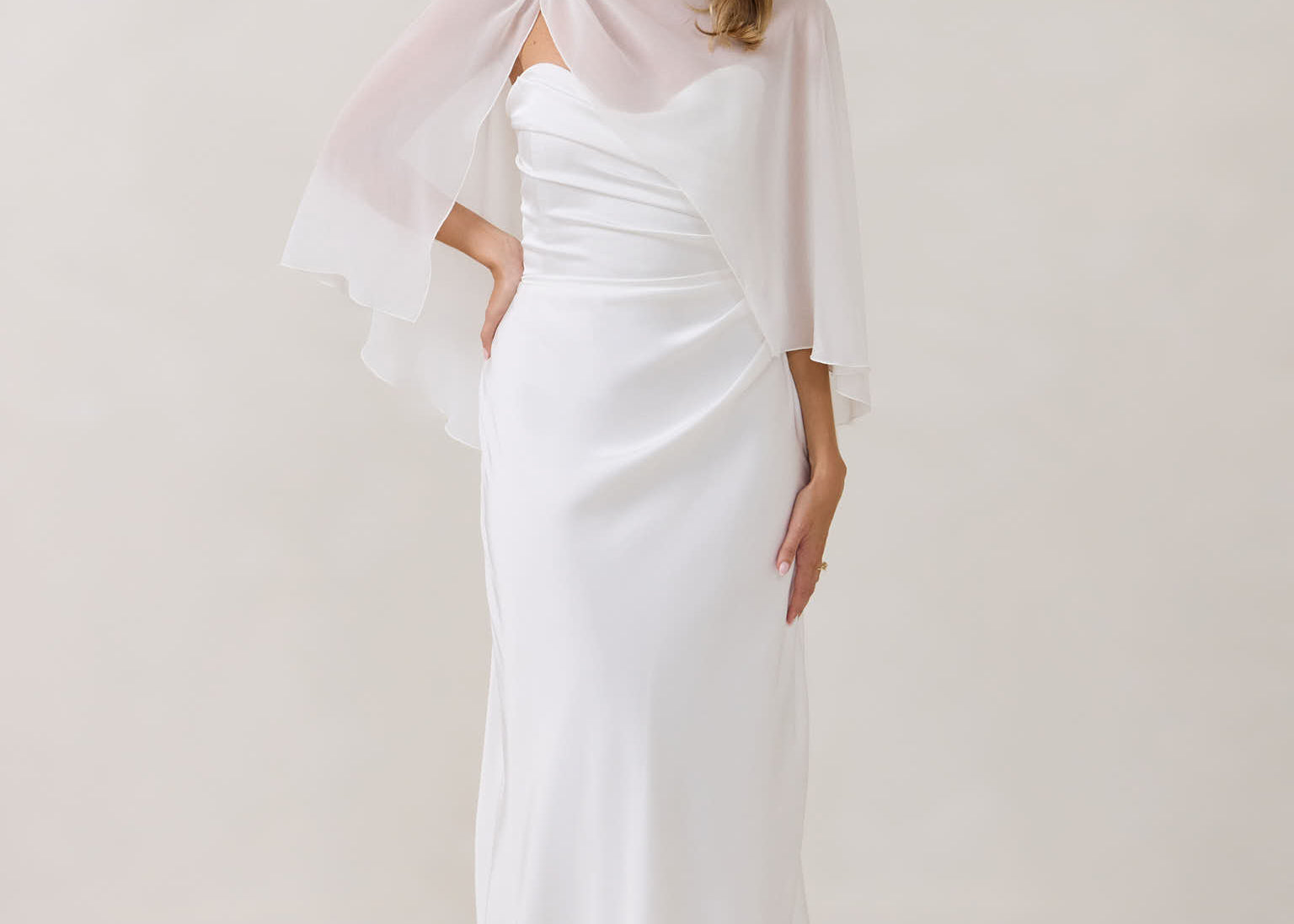 A woman is standing against a white backdrop. She's wearing a white bridal dress with a chiffon cape over her shoulders. The cape is gathered slightly on the left side and is nearly fingertip length. This is the Cleo Cape.