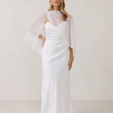 A woman is standing against a white backdrop. She's wearing a white bridal dress with a chiffon cape over her shoulders. The cape is gathered slightly on the left side and is nearly fingertip length. This is the Cleo Cape.