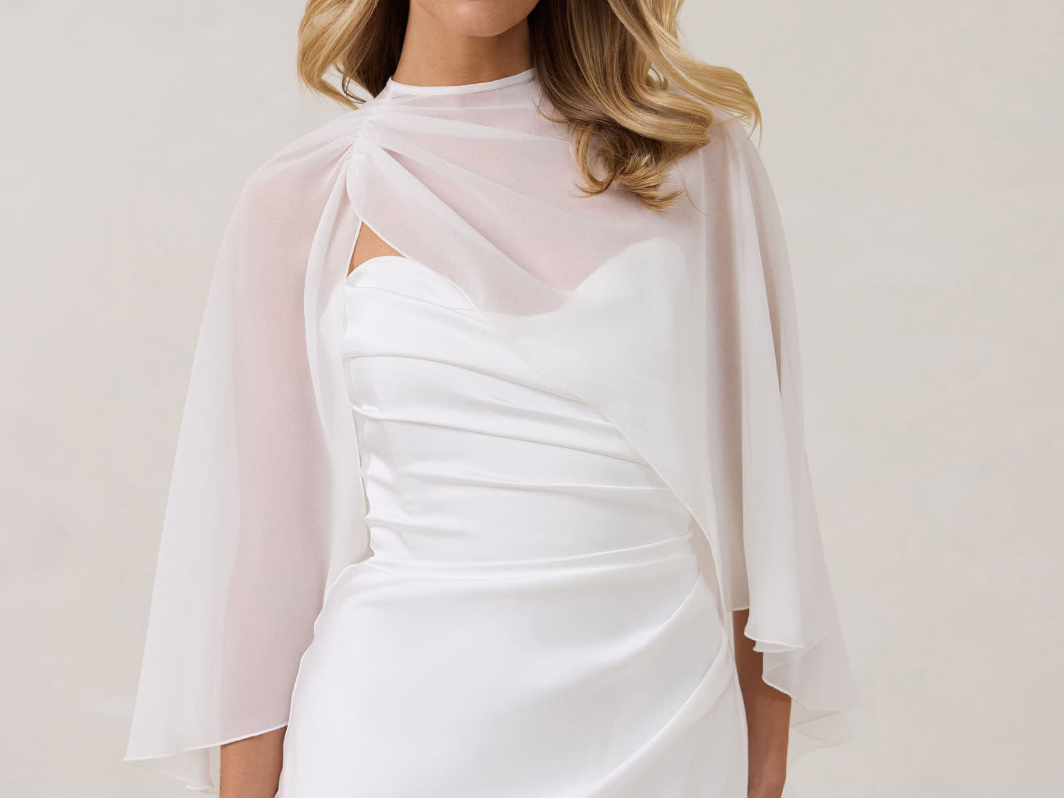 A woman is standing against a white backdrop. She's wearing a white bridal dress with a chiffon cape over her shoulders. The cape is gathered slightly on the left side and is nearly fingertip length. This is the Cleo Cape.