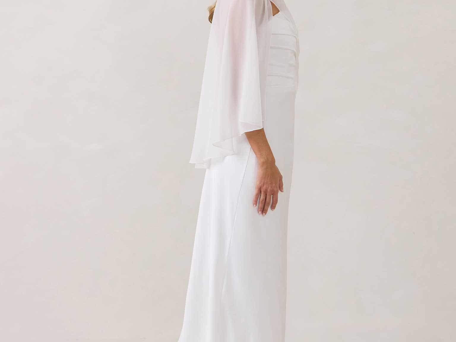 A woman is standing against a white backdrop. She's wearing a white bridal dress with a chiffon cape over her shoulders. The cape is gathered slightly on the left side and is nearly fingertip length. This is the Cleo Cape.