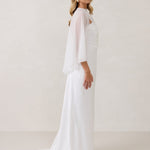 A woman is standing against a white backdrop. She's wearing a white bridal dress with a chiffon cape over her shoulders. The cape is gathered slightly on the left side and is nearly fingertip length. This is the Cleo Cape.