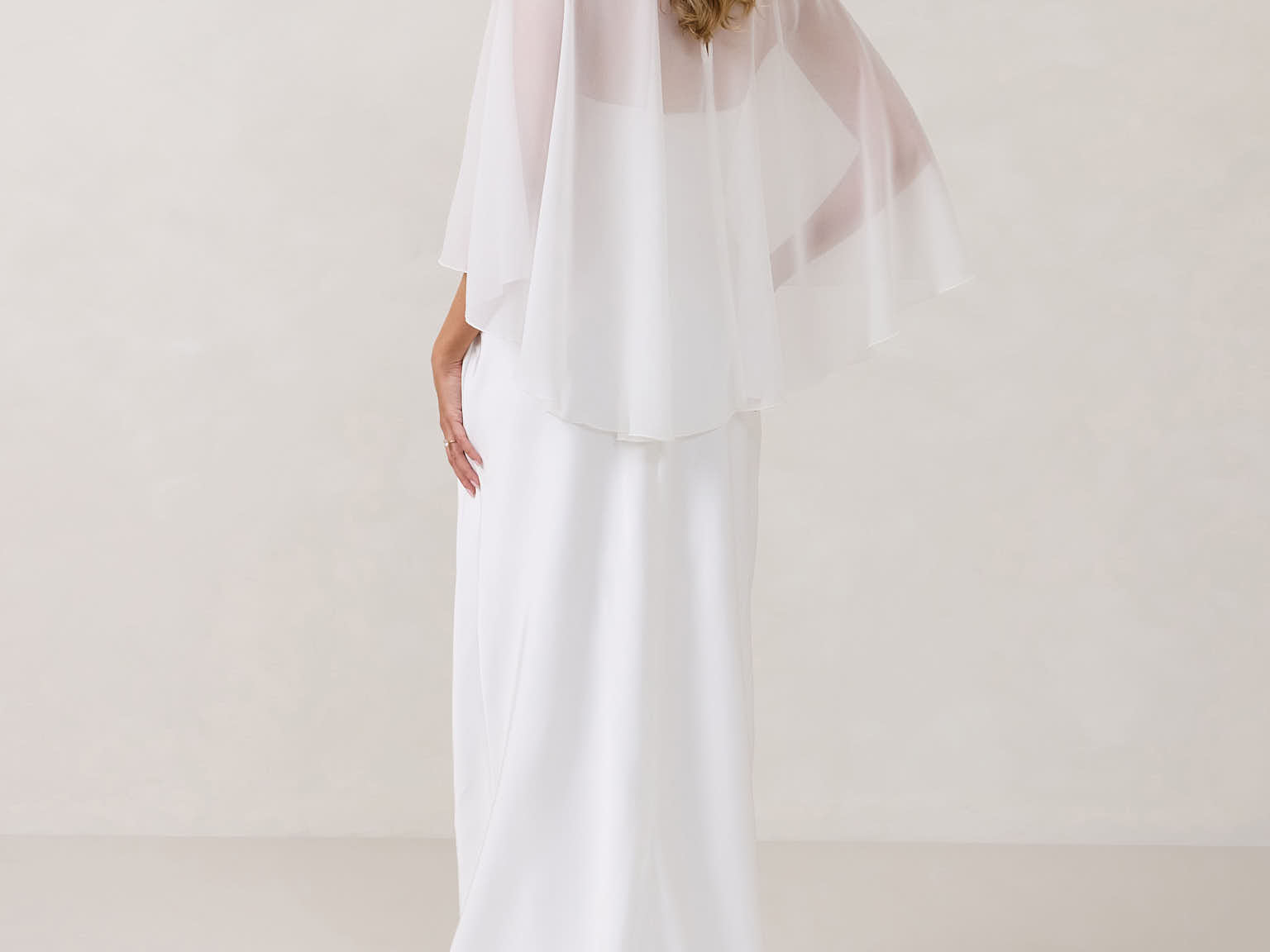 A woman is standing against a white backdrop facing the back. She's wearing a white bridal dress with a chiffon cape over her shoulders. The cape is gathered slightly on the left side and is nearly fingertip length. This is the Cleo Cape.