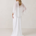 A woman is standing against a white backdrop facing the back. She's wearing a white bridal dress with a chiffon cape over her shoulders. The cape is gathered slightly on the left side and is nearly fingertip length. This is the Cleo Cape.