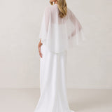 A woman is standing against a white backdrop facing the back. She's wearing a white bridal dress with a chiffon cape over her shoulders. The cape is gathered slightly on the left side and is nearly fingertip length. This is the Cleo Cape.