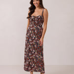 A woman with long dark hair is standing infront of a floral display. She's wearing a floral patterned fitted dress. The dress is fitted through the bodice and has a keyhole detail with a tie at the bust. This spring dress is tea-length and has shirring at the back for added comfort. This is the Decker Dress in Isabel. 