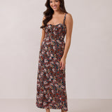 A woman with long dark hair is standing infront of a floral display. She's wearing a floral patterned fitted dress. The dress is fitted through the bodice and has a keyhole detail with a tie at the bust. This spring dress is tea-length and has shirring at the back for added comfort. This is the Decker Dress in Isabel. 