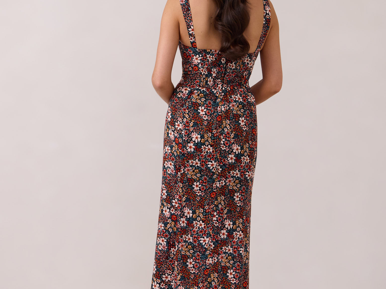 A woman with long dark hair is standing infront of a floral display. She's wearing a floral patterned fitted dress. The dress is fitted through the bodice and has a keyhole detail with a tie at the bust. This spring dress is tea-length and has shirring at the back for added comfort. This is the Decker Dress in Isabel. 