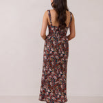 A woman with long dark hair is standing infront of a floral display. She's wearing a floral patterned fitted dress. The dress is fitted through the bodice and has a keyhole detail with a tie at the bust. This spring dress is tea-length and has shirring at the back for added comfort. This is the Decker Dress in Isabel. 