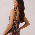 A woman with long dark hair is standing infront of a floral display. She's wearing a floral patterned fitted dress. The dress is fitted through the bodice and has a keyhole detail with a tie at the bust. This spring dress is tea-length and has shirring at the back for added comfort. This is the Decker Dress in Isabel. 