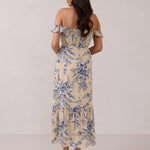 A woman is standing against a white backdrop facing the back. She's wearing a tea-length cream dress with a delicate blue floral print. The dress has a ruffle detail around the neckline that can be worn on or off the shoulder. The skirt has a slit up to the thigh and a ruffle detail around the hem. This is the Renfrew Dress in Ayla. 